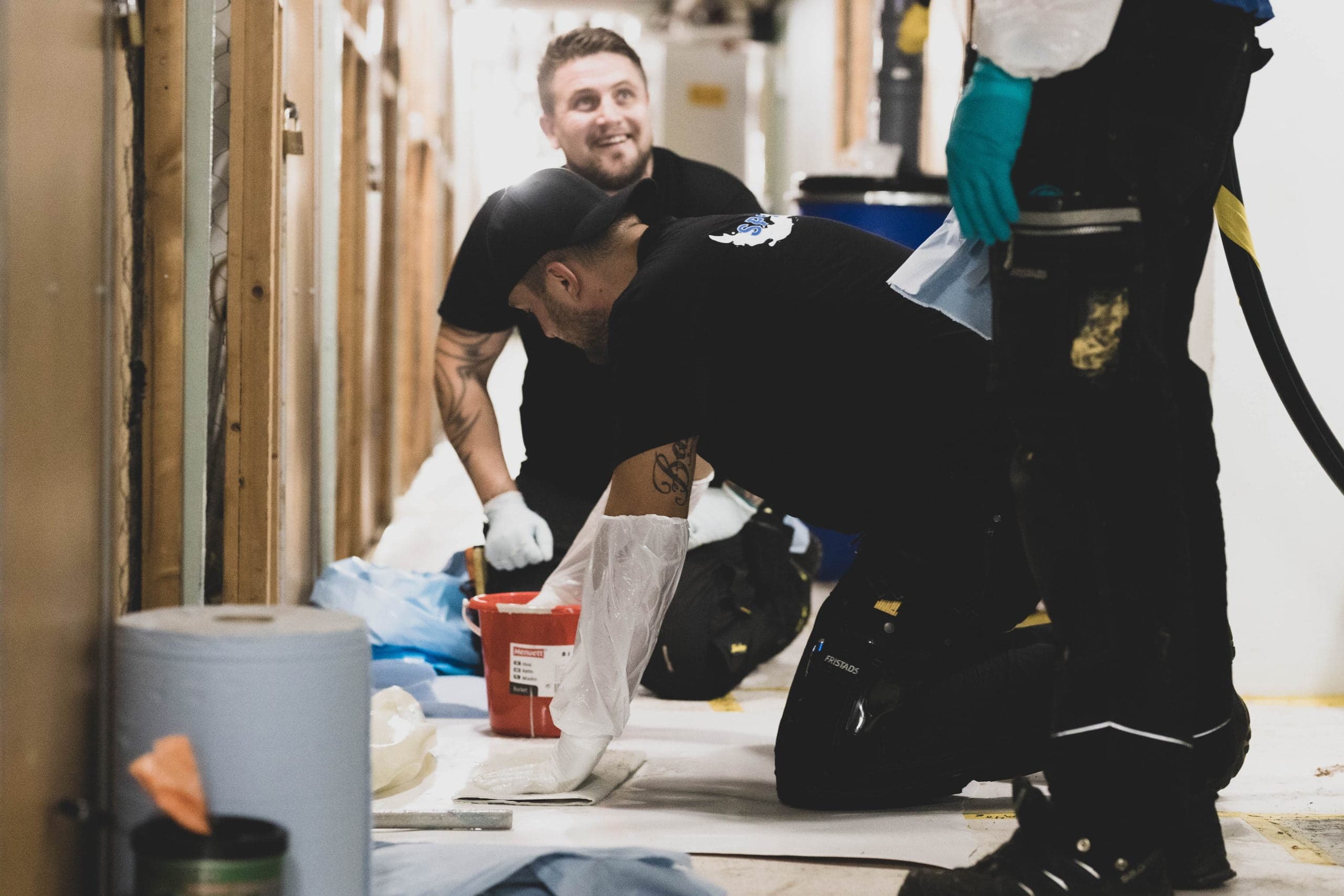 Spoltec-arbetare i handskar, en på knä med en hink, den andra ler bakom.